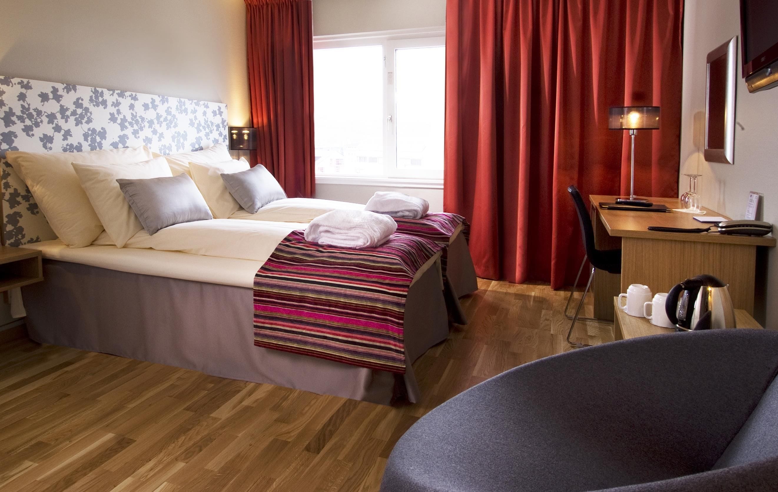 Een hotelkamer met een bed, een bureau, een stoel, een lamp en een raam met gordijnen.