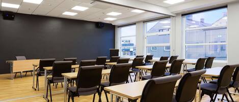 A classroom with rows of desks and chairs, large windows, and a projector on the ceiling.