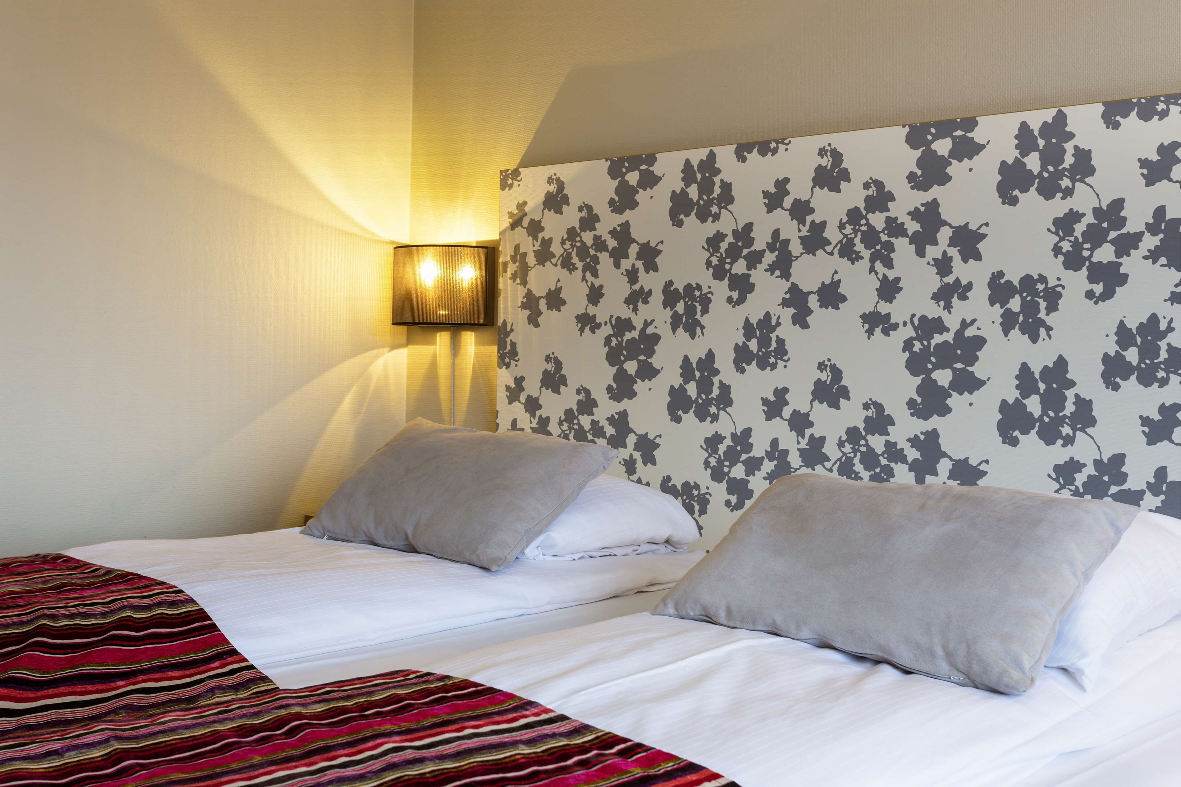 Ein Hotelzimmer mit zwei Betten, einer gestreiften Decke, floralem Wandpapier und einer Wandleuchte.