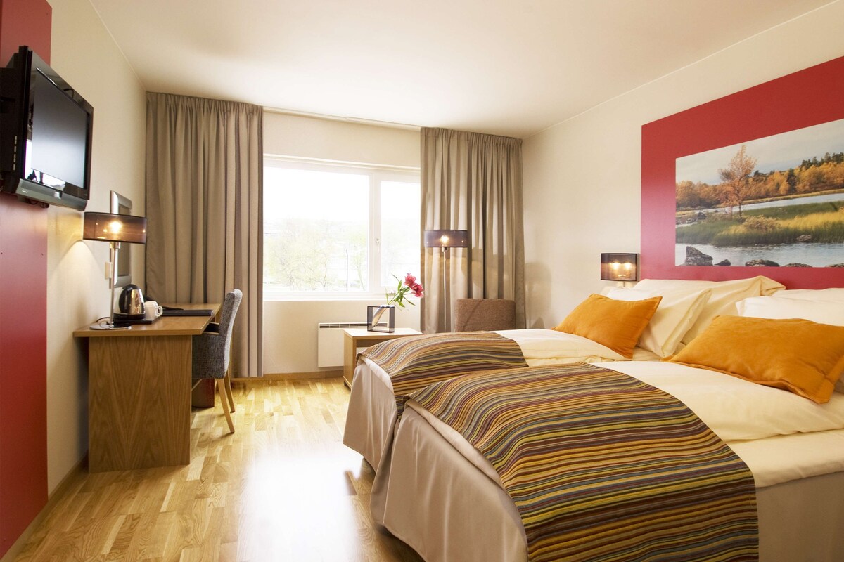 Una habitación de hotel con cama, escritorio, silla, televisor y ventana con cortinas.