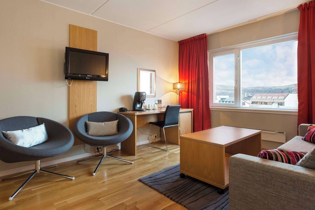 Una habitación de hotel moderna con un escritorio de madera, dos sillas, un sofá, un televisor y un ventanal con vistas a edificios.