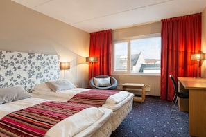 Une chambre d’hôtel avec deux lits, un bureau, une chaise, une fenêtre avec des rideaux et une lampe.