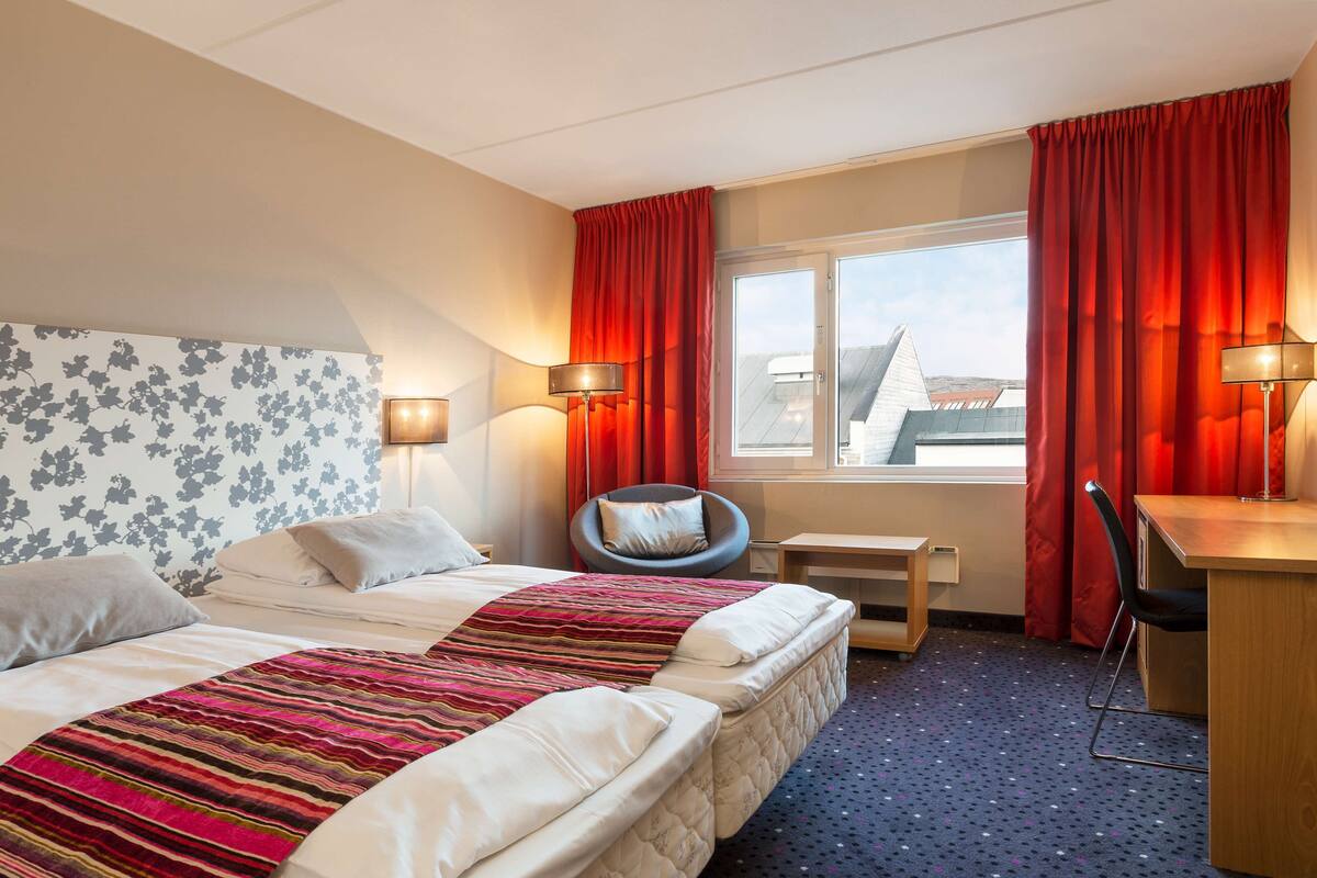 Habitación de hotel con dos camas, un escritorio, una silla, un ventanal con cortinas y una lámpara.