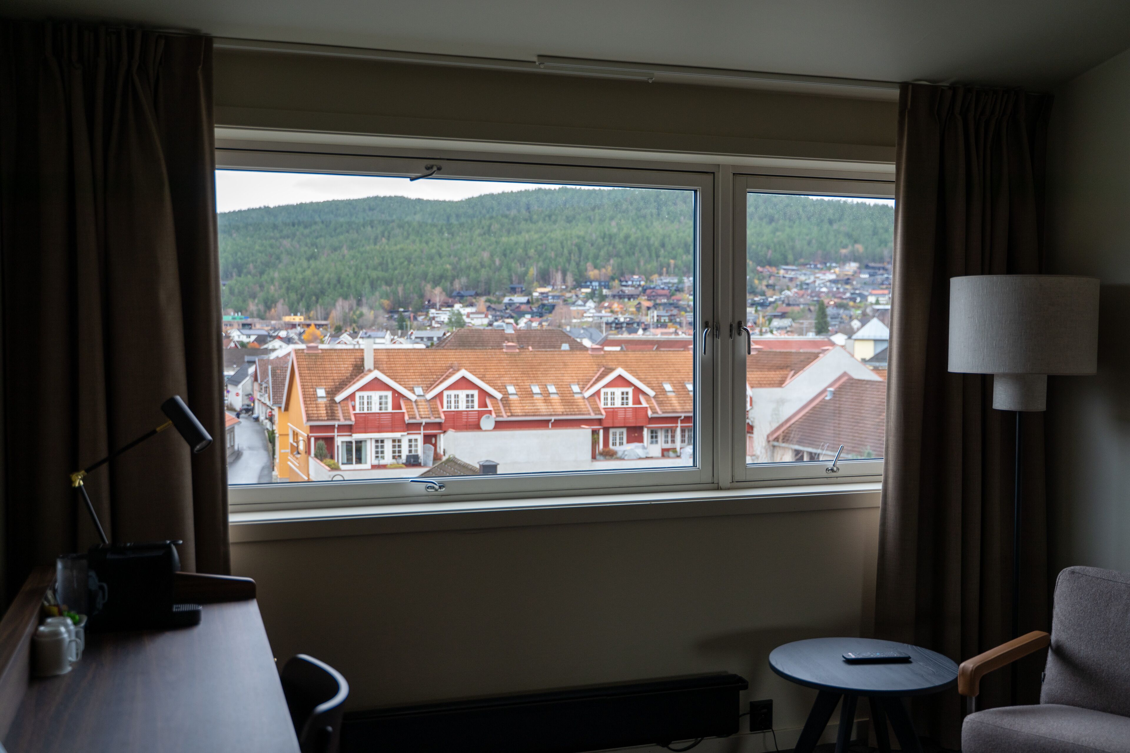 A hotel room with a large window offering a view of a town and forested hills.