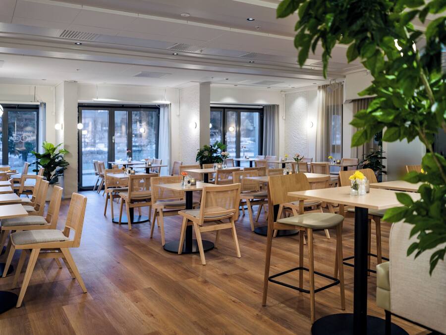 Un comedor amplio con mesas y sillas de madera, ventanas grandes y plantas de interior.