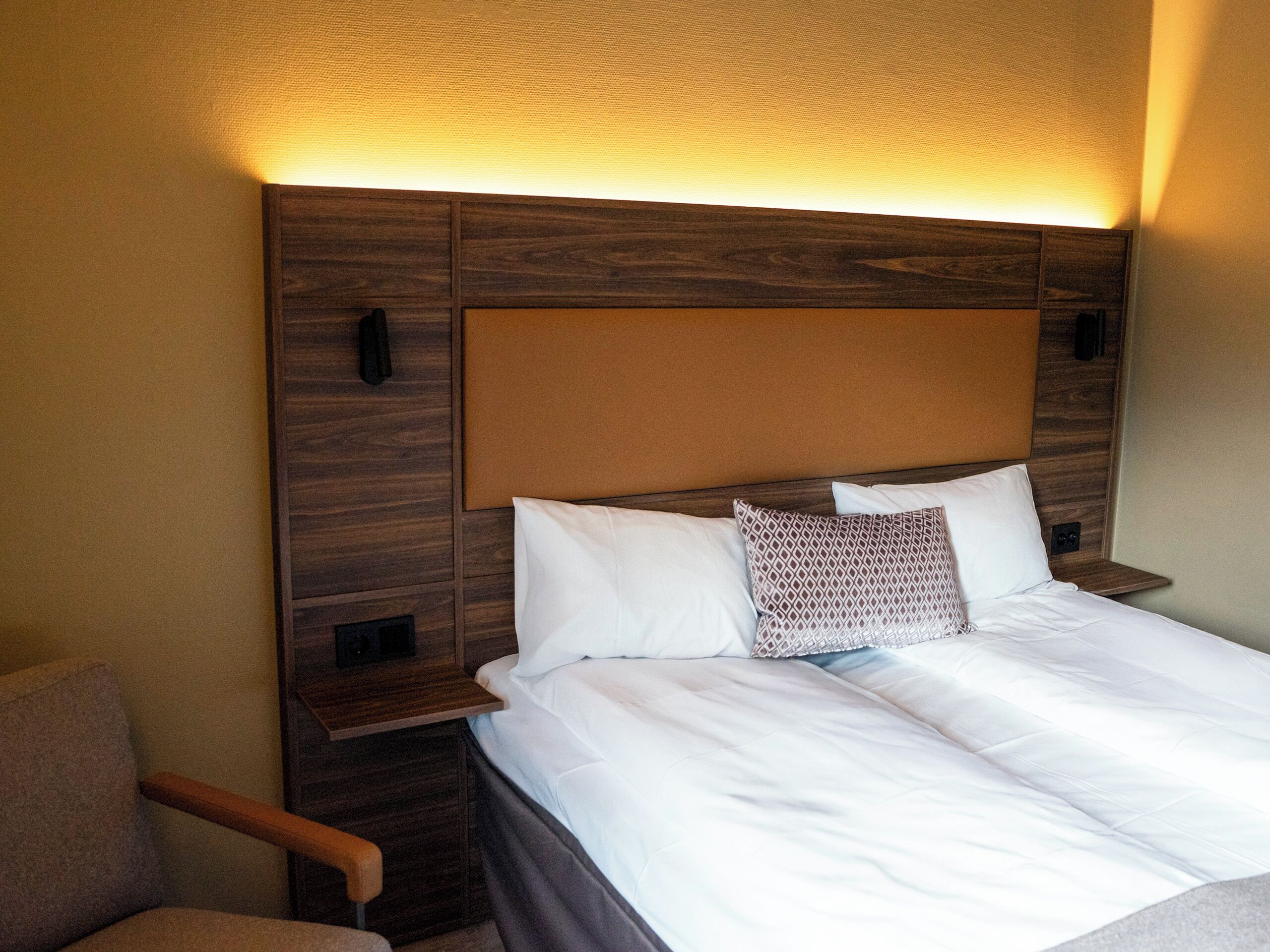 A hotel room with a wooden headboard, a bed with white linens, a patterned pillow, and a nightstand with a phone.