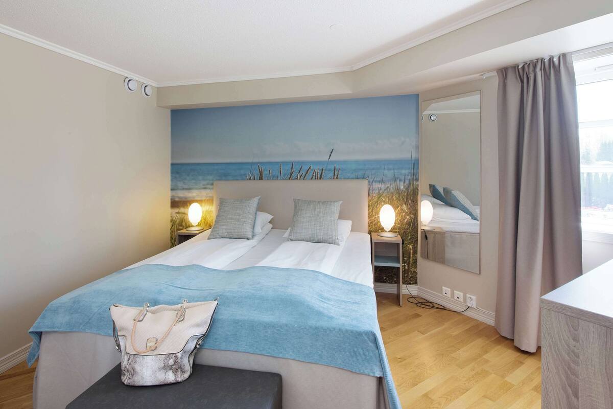 A double bed with a blue blanket, a bedside table with a lamp, and a view of the sea through a window.