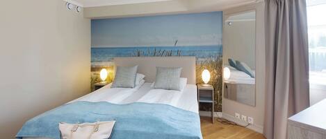 A double bed with a blue blanket, a bedside table with a lamp, and a view of the sea through a window.