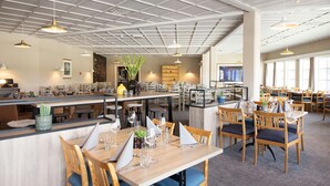 Un restaurante con mesas y sillas de madera, una zona de bar y vistas a la ciudad a través de amplios ventanales.