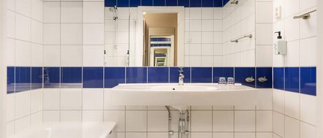 A bathroom with a white sink, blue-tiled walls, a bathtub, and a mirror reflecting a hallway.