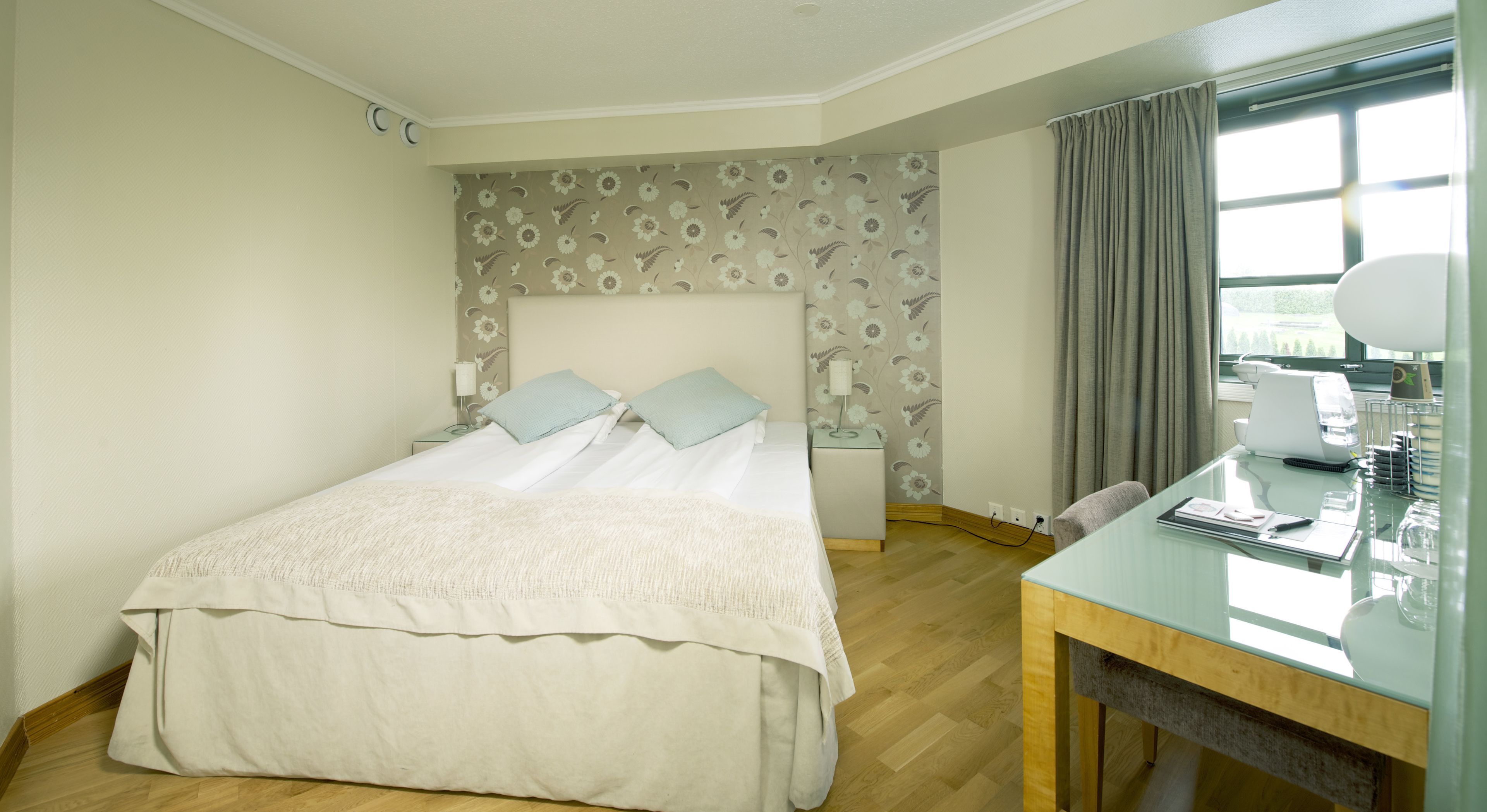 A hotel room with a bed, a desk with a chair, a window with curtains, and a wall with floral wallpaper.
