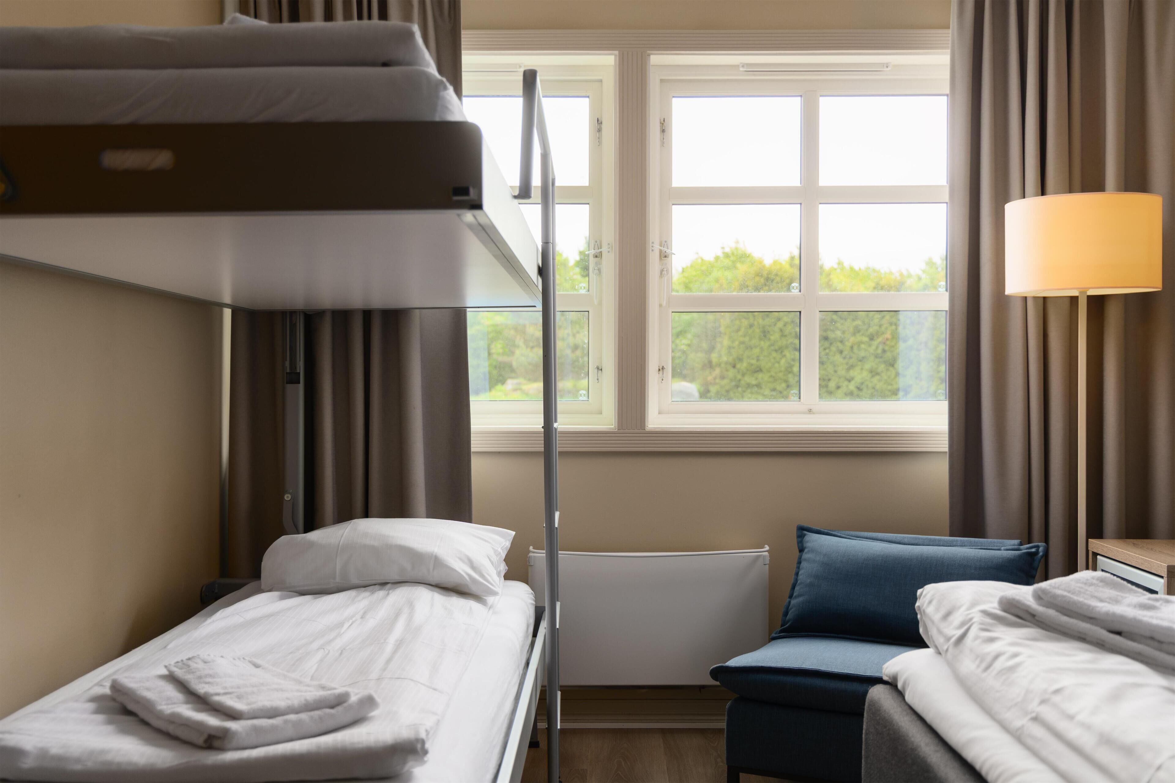 Una habitación de hotel con una cama nido, una cama individual, un escritorio con lámpara y una ventana con cortinas.