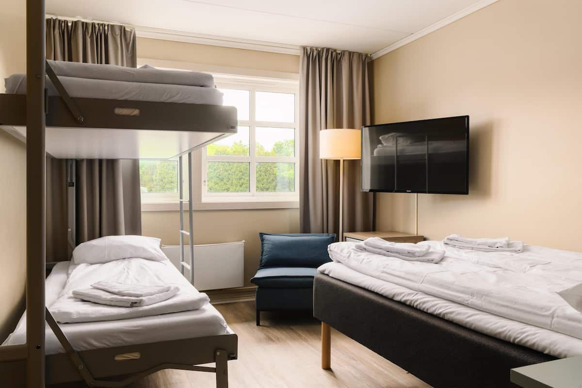 A hotel room with a bunk bed, a single bed, a desk, a chair, a TV, and a window with curtains.