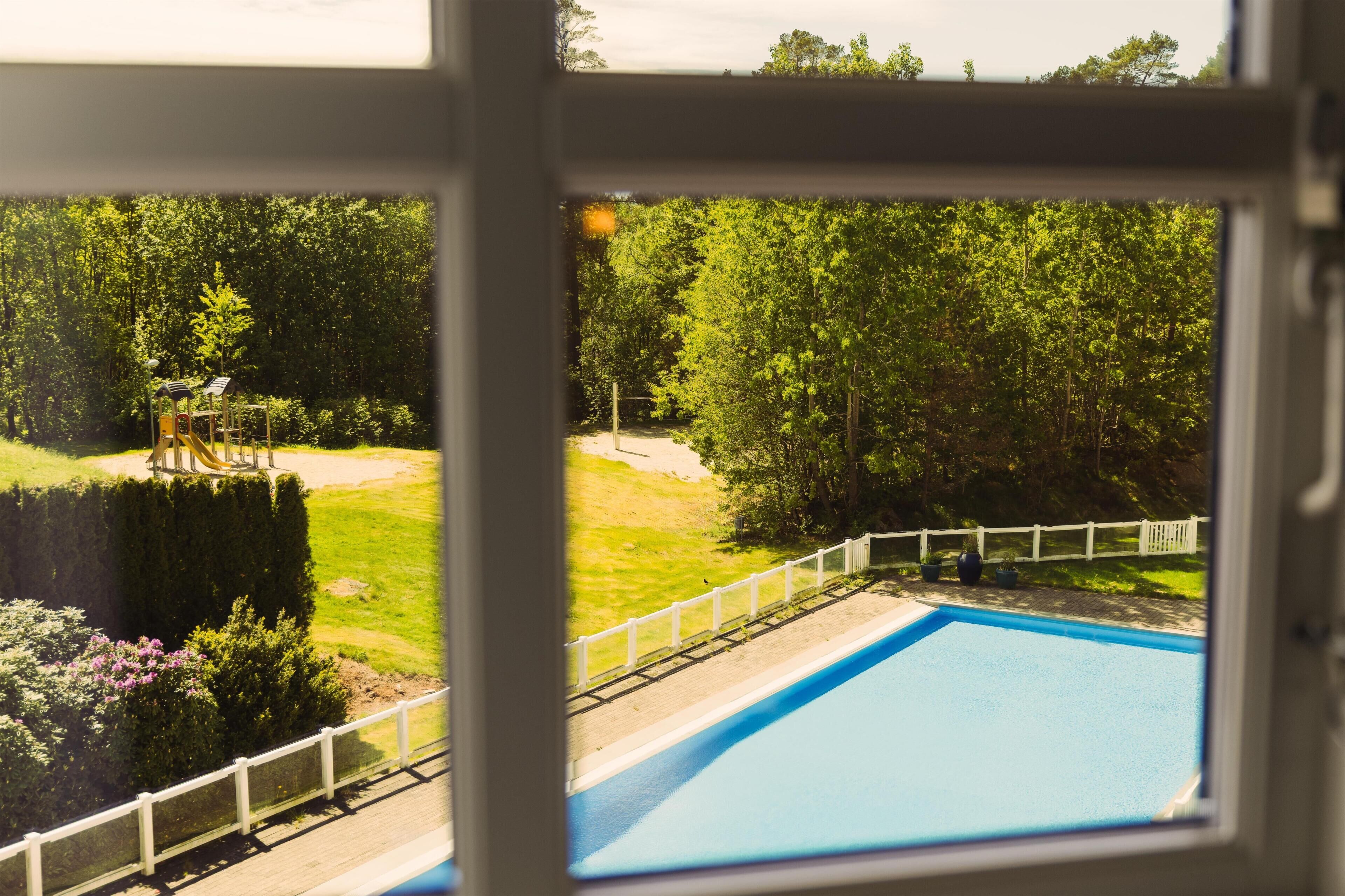 Vista a través de una ventana de una piscina, rodeada por una cerca blanca, con un parque infantil y árboles frondosos al fondo.