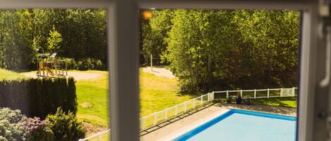 Vista a través de una ventana de una piscina, rodeada por una cerca blanca, con un parque infantil y árboles frondosos al fondo.