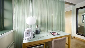 A hotel room with a wooden desk, a white lamp, a coffee maker, and a glass table.