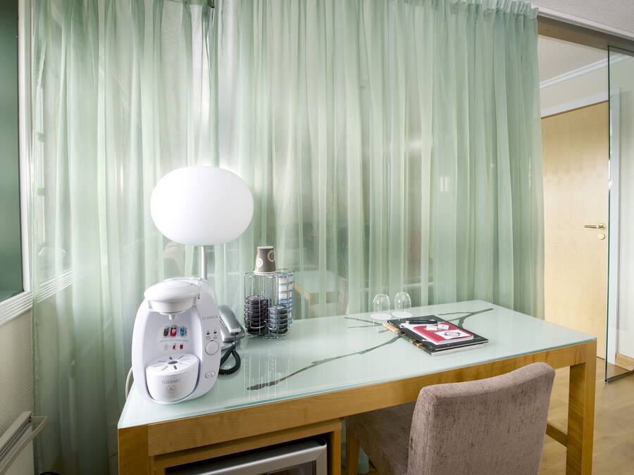A hotel room with a wooden desk, a white lamp, a coffee maker, and a glass table.