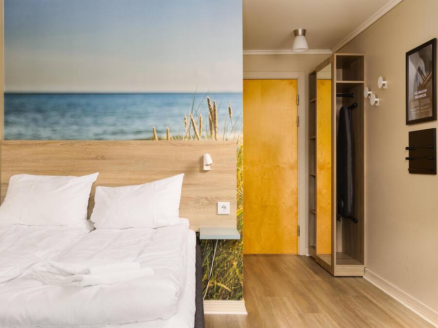 Un dormitorio con una cama, una mesita de noche, una lámpara de pared, un armario y una puerta que da a otra habitación.