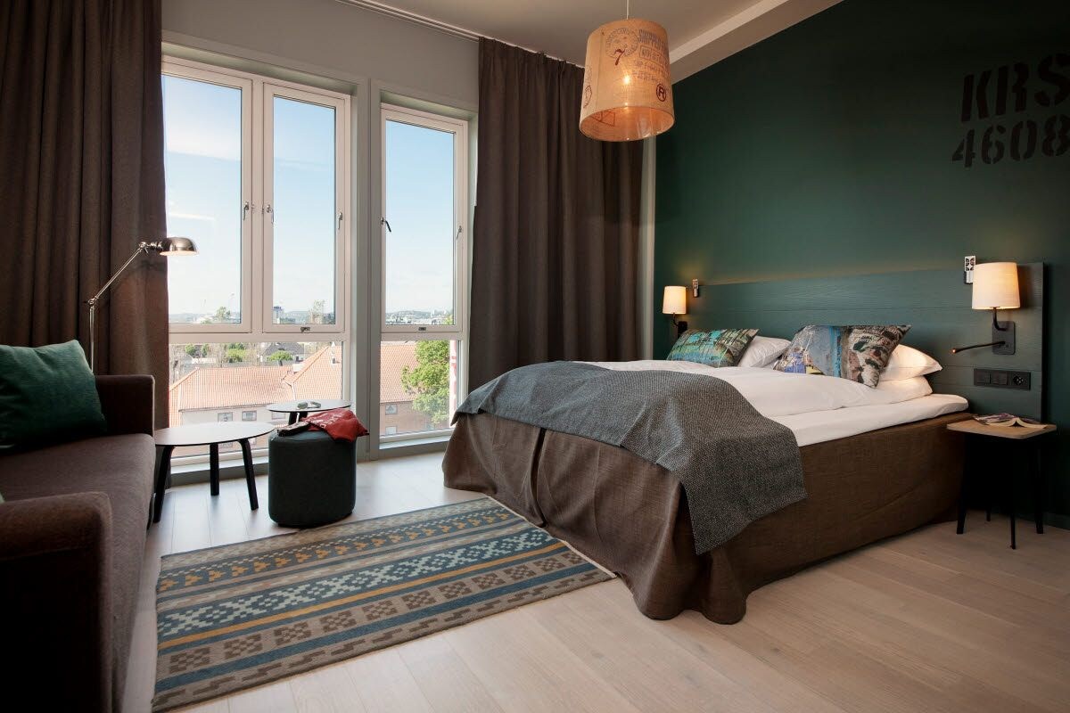 A bedroom with a large bed, a view through a window, a sofa, a lamp, and a wall-mounted light.