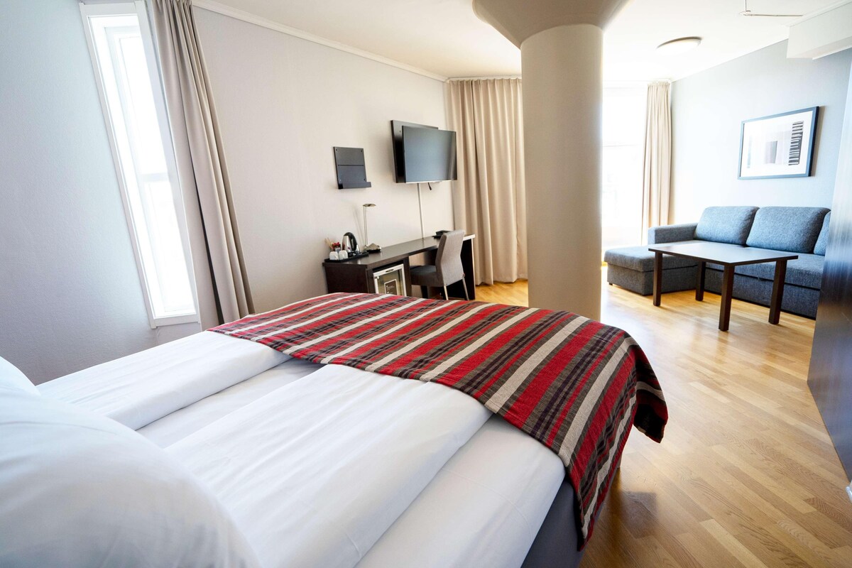 A hotel room with a large bed, a desk with a chair, a TV, and a sofa.