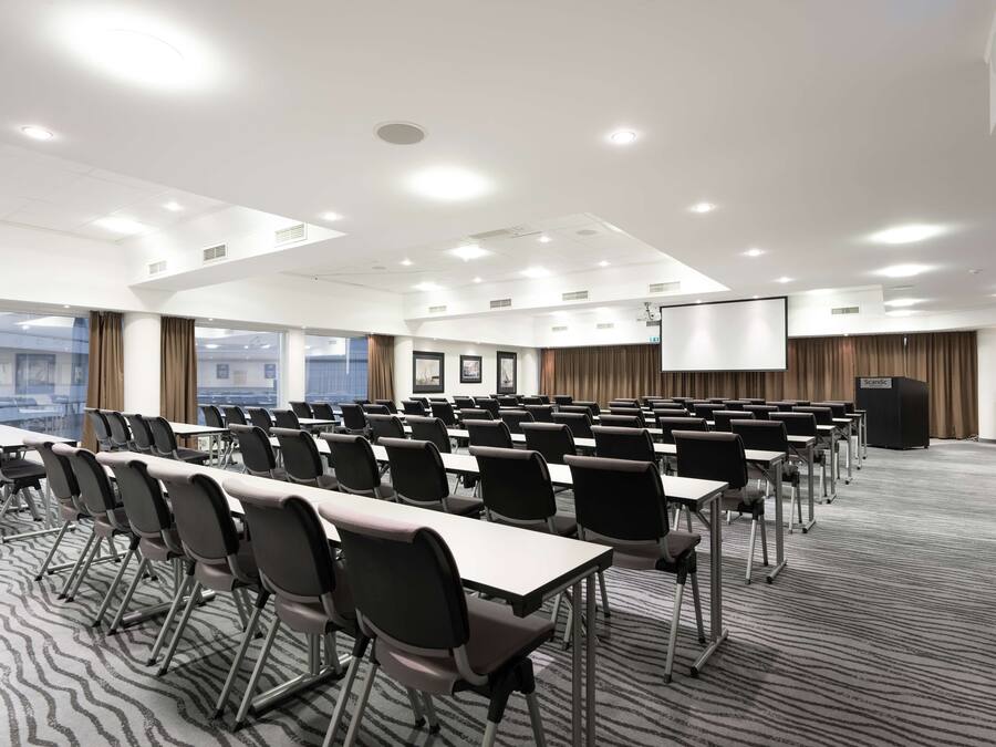 Une salle de conférence avec des rangées de tables et de chaises, un écran de projection et un pupitre.