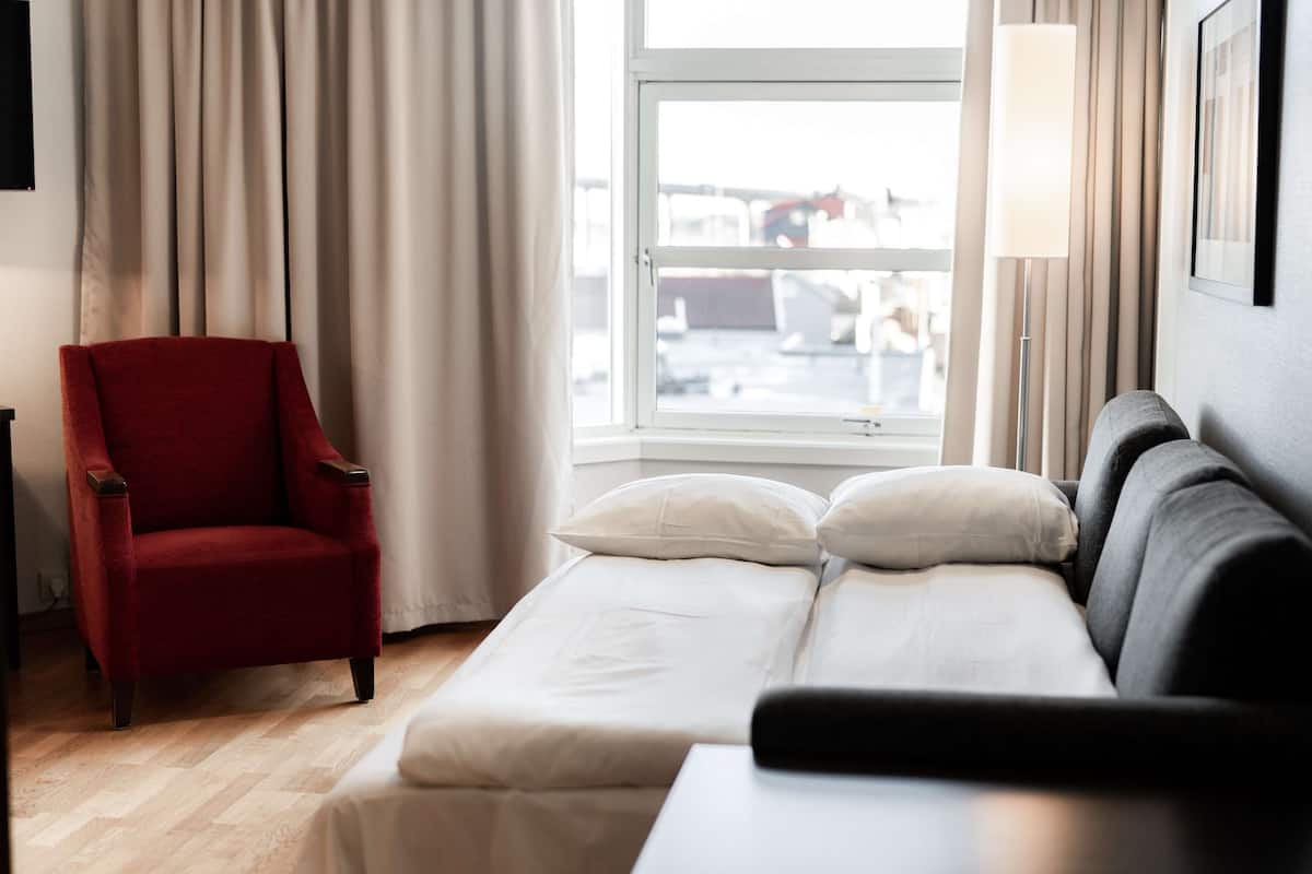 A hotel room with a red armchair, a sofa, a bed with white bedding, a window with curtains, and a floor lamp.