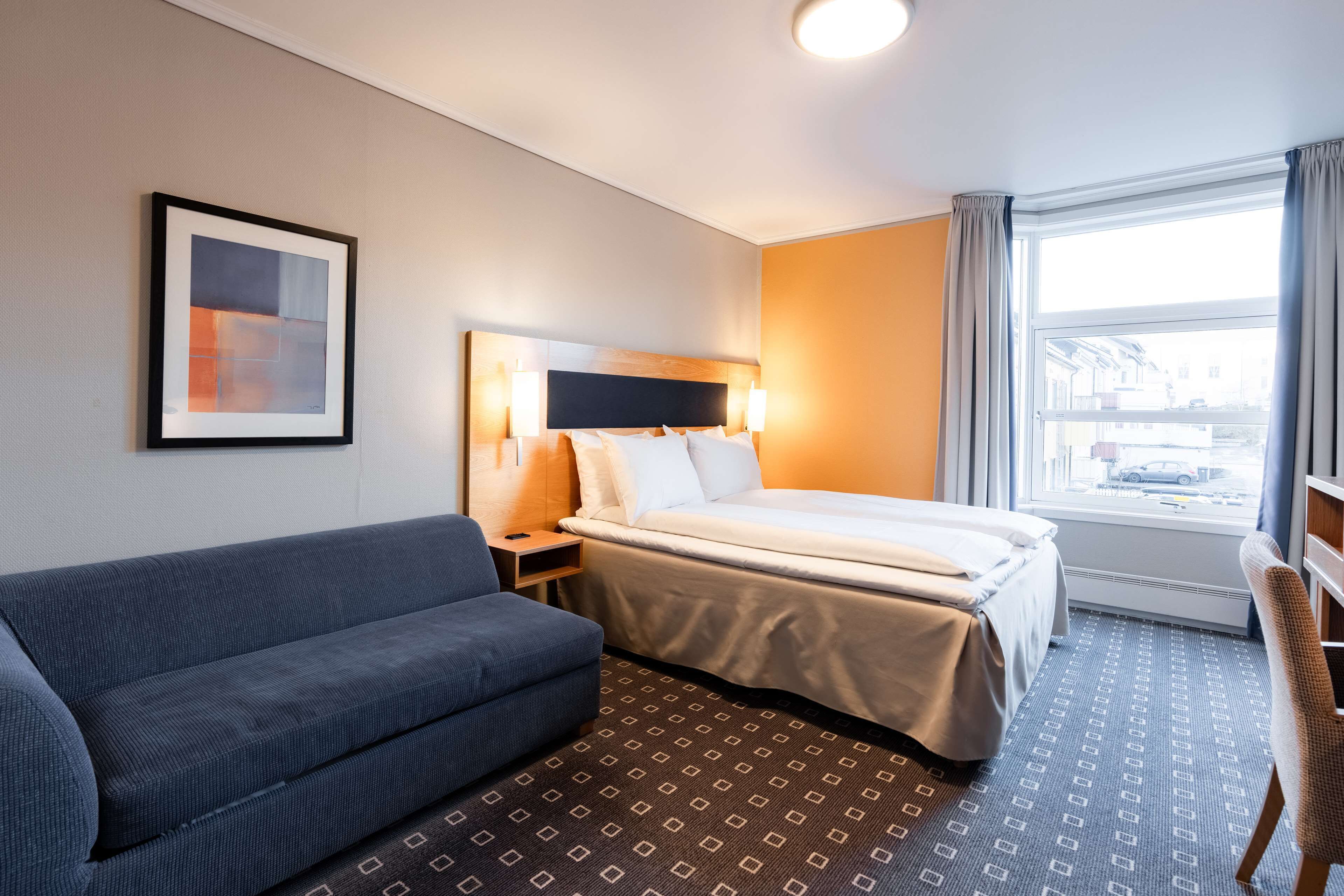 A hotel room with a bed, a sofa, a nightstand, a lamp, and a window with a view of the street.
