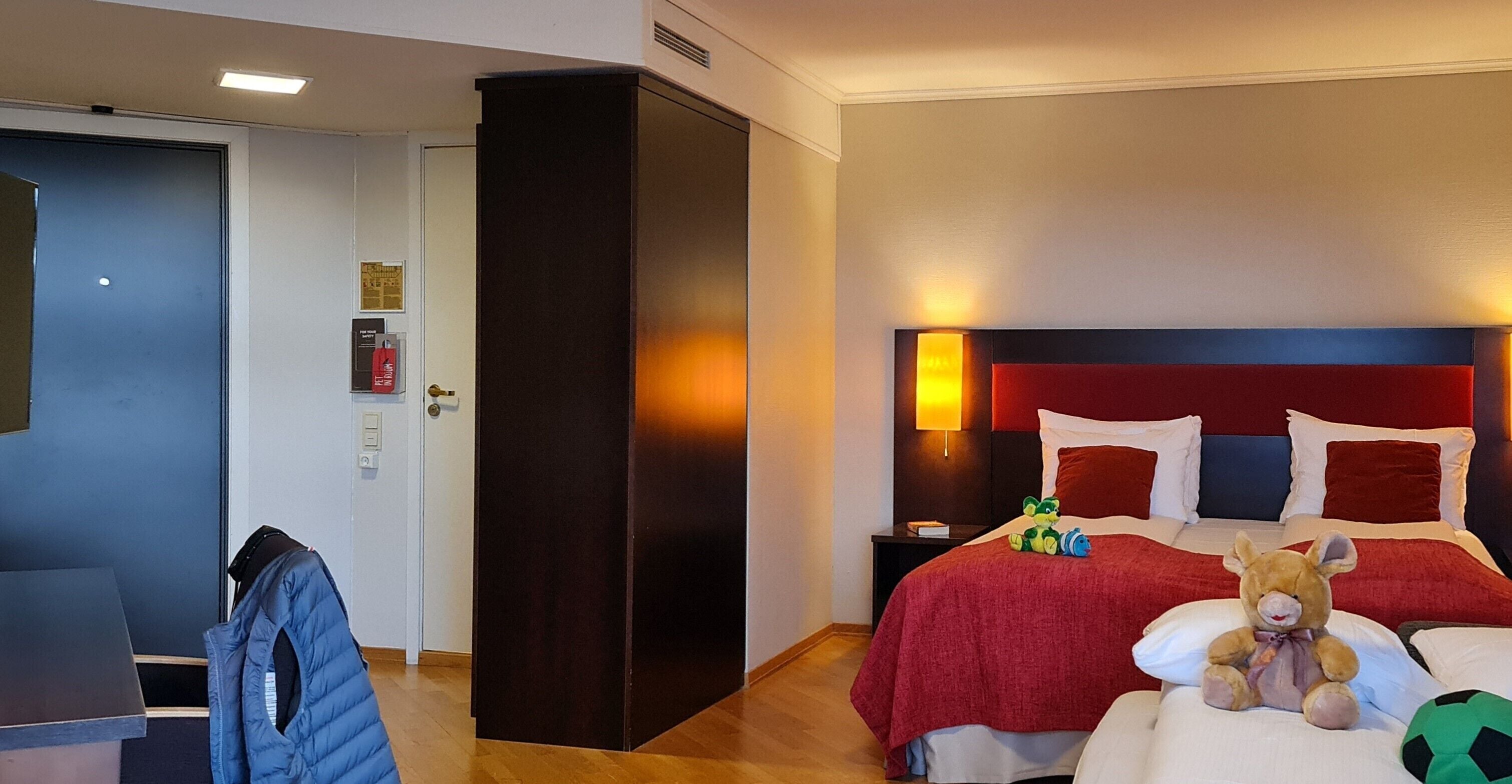 A hotel room with a bed, a nightstand, a lamp, a teddy bear, and a jacket on a chair.