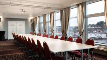 Un salón de conferencias con una larga mesa y sillas rojas, ventanas amplias con cortinas y vistas a la ciudad.