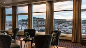 Quarto de hotel com grandes janelas, vista para a paisagem urbana e área de estar com cadeiras e uma mesa.