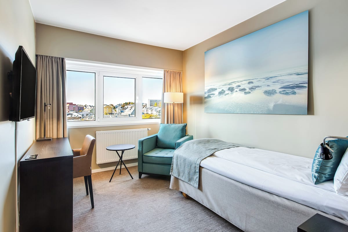 Une chambre d’hôtel avec un lit, un bureau, une chaise, une fenêtre donnant sur des immeubles et un tableau accroché au mur.