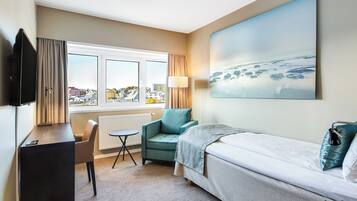 Une chambre d’hôtel avec un lit, un bureau, une chaise, une fenêtre donnant sur des immeubles et un tableau accroché au mur.