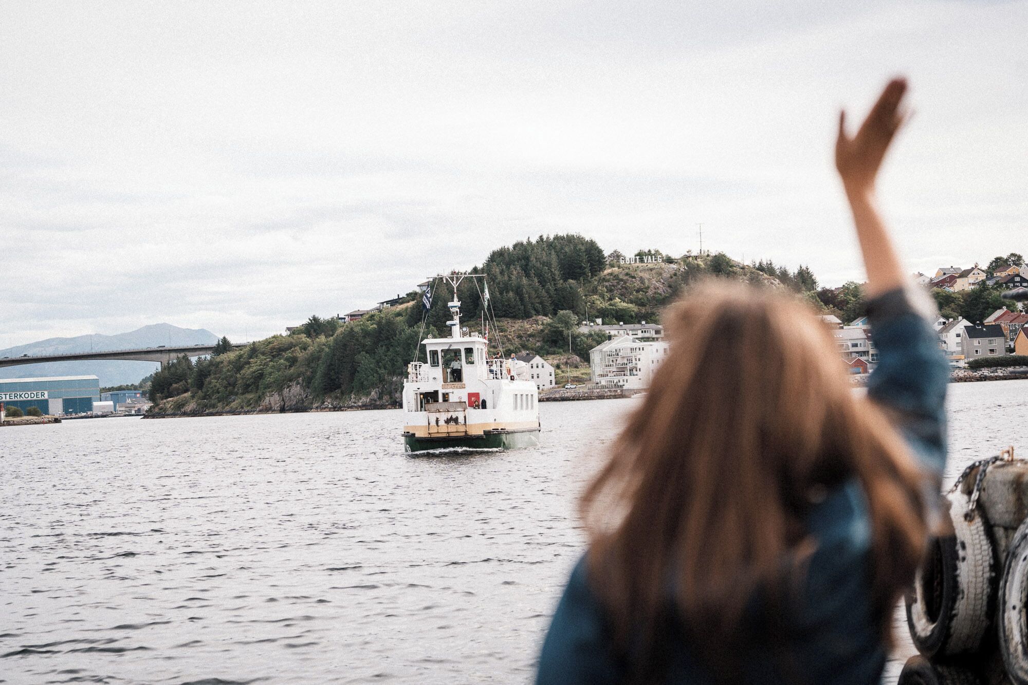 En person med hævede arme står og ser på en båd på vandet, med bygninger i baggrunden.