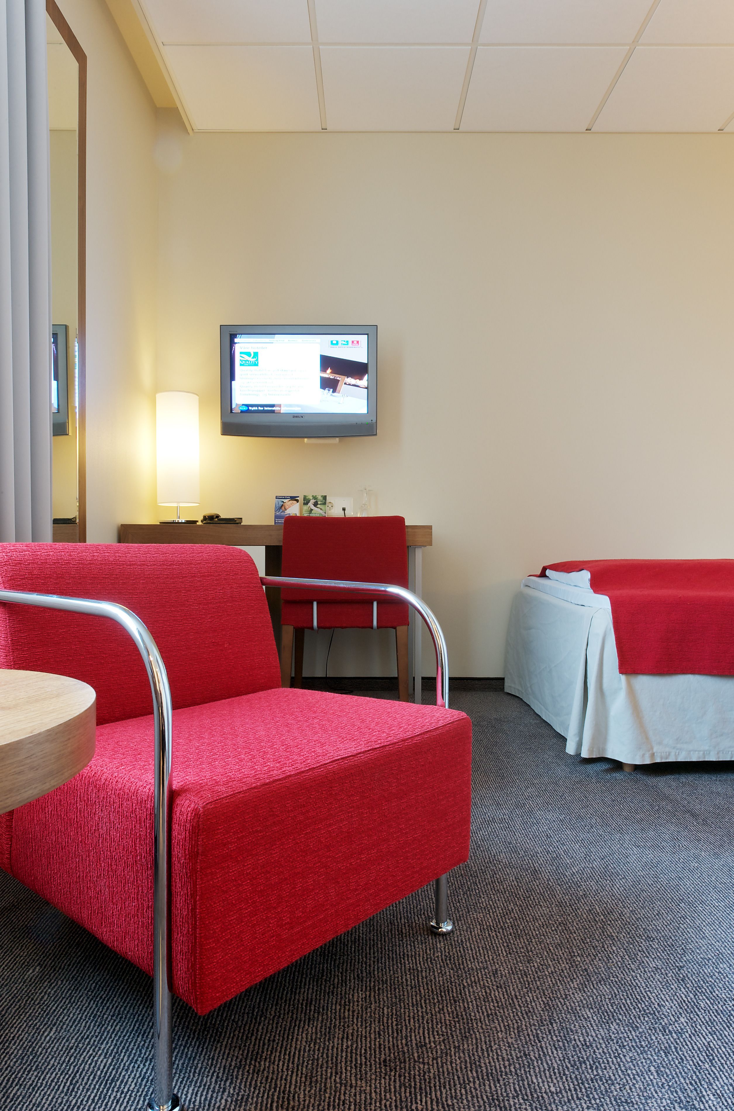 Una habitación de hotel con un sillón rojo, una mesita pequeña, un escritorio con lámpara, una cama con manta roja y un televisor en la pared.