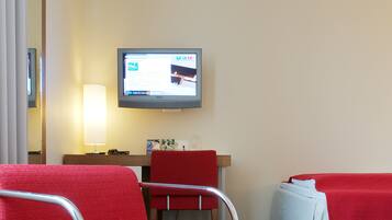 Una habitación de hotel con un sillón rojo, una mesita pequeña, un escritorio con lámpara, una cama con manta roja y un televisor en la pared.
