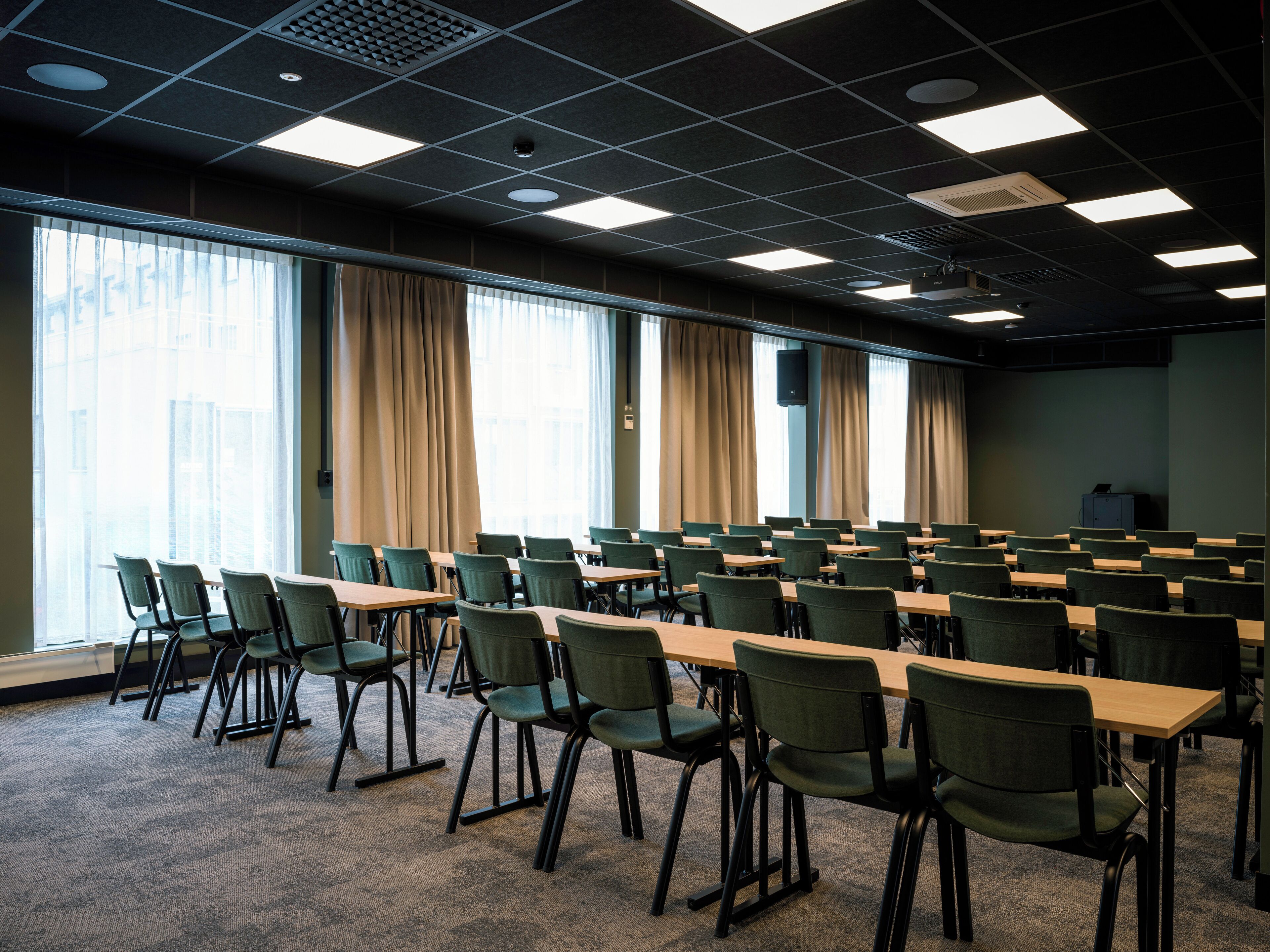 Un salón de conferencias con filas de mesas y sillas, ventanas amplias con cortinas e iluminación en el techo.