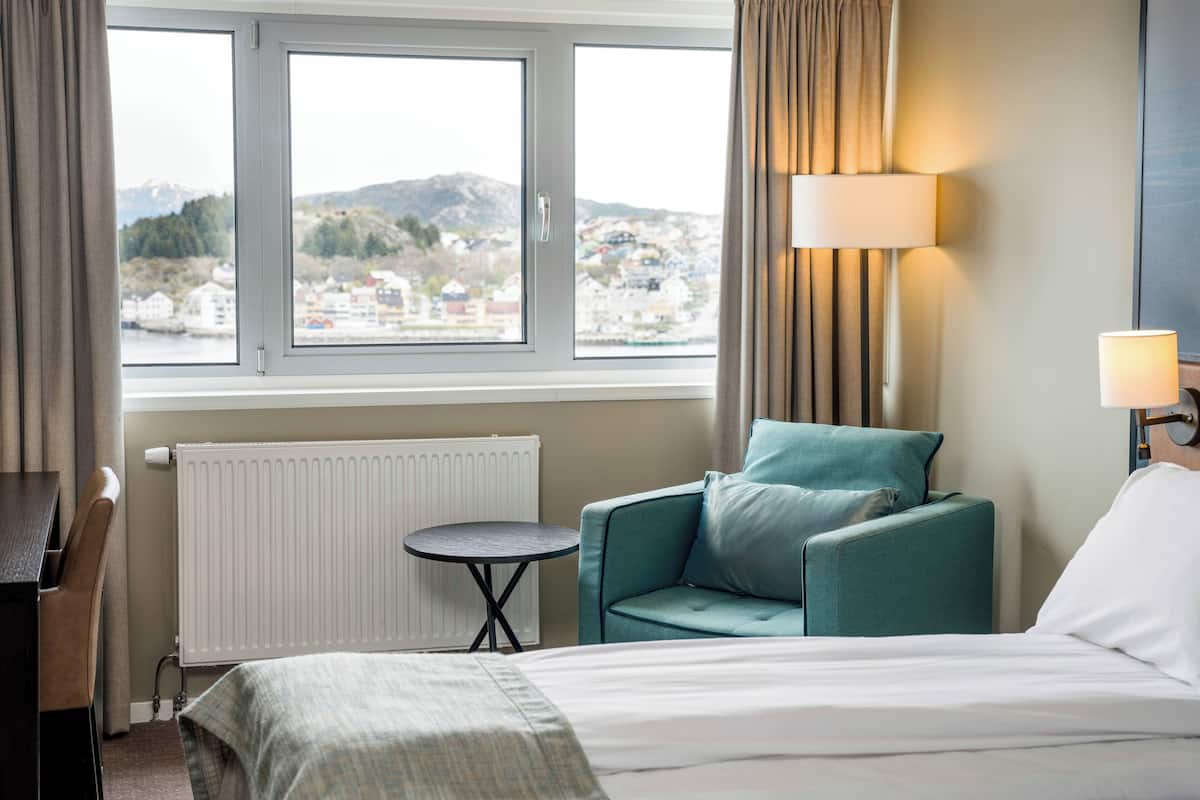 Une chambre d’hôtel avec un lit, une chaise, une petite table, une lampe et une vue sur le paysage urbain, visible à travers de grandes fenêtres.