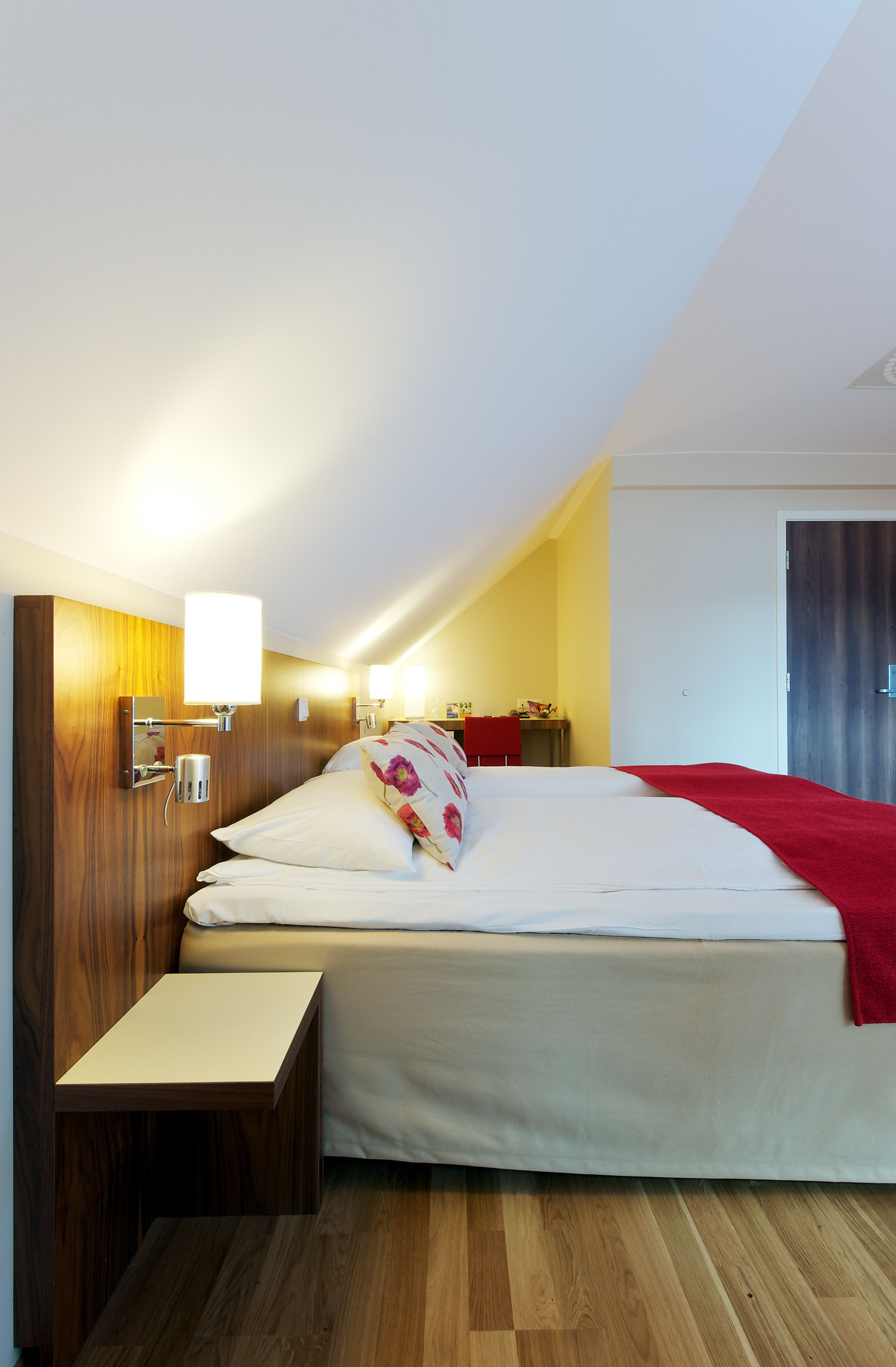 A hotel room with a wooden headboard, a bed with white bedding and a red blanket, a bedside table, and a wall-mounted lamp.