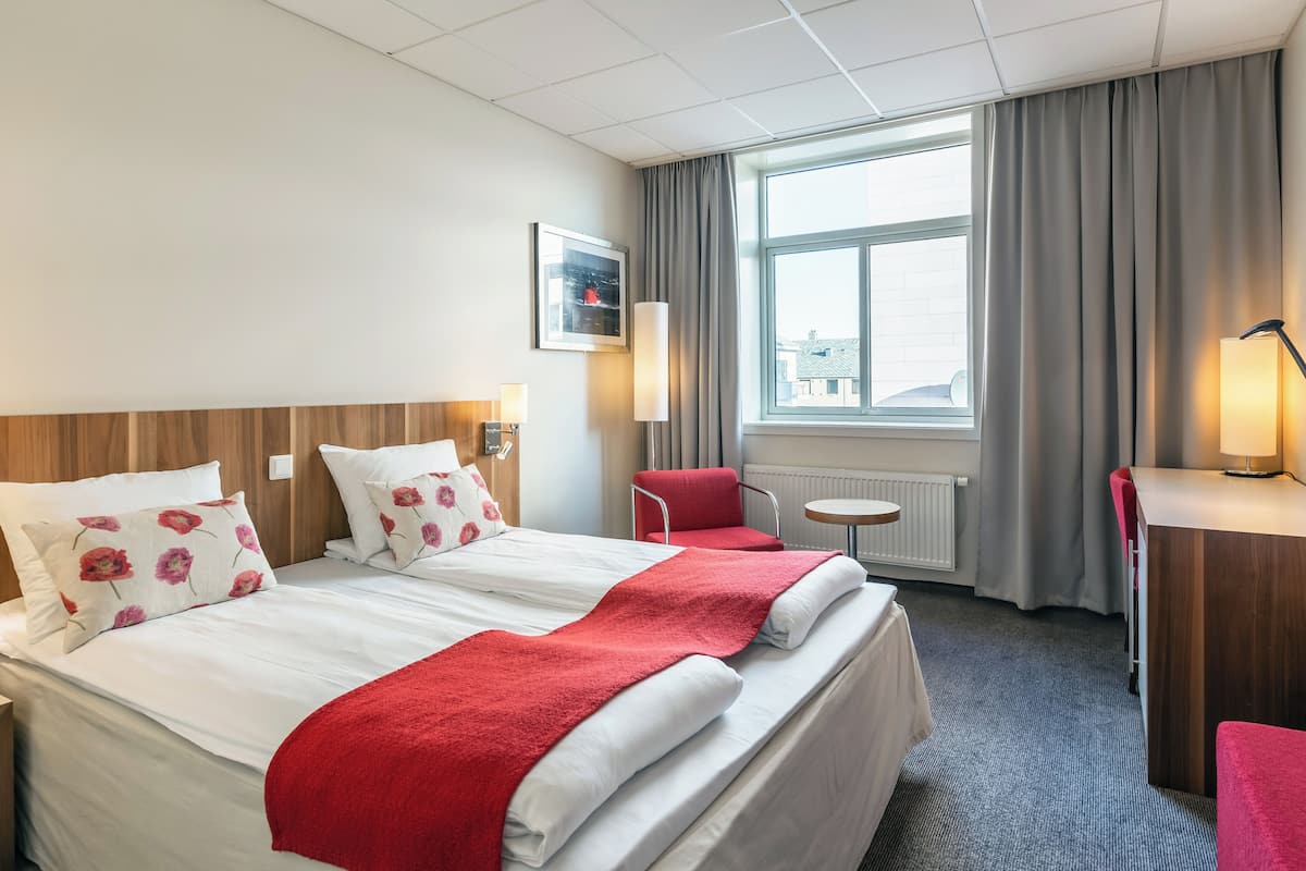 Une chambre d’hôtel avec un grand lit, deux tables de chevet, une chaise, un bureau et une fenêtre avec des rideaux.