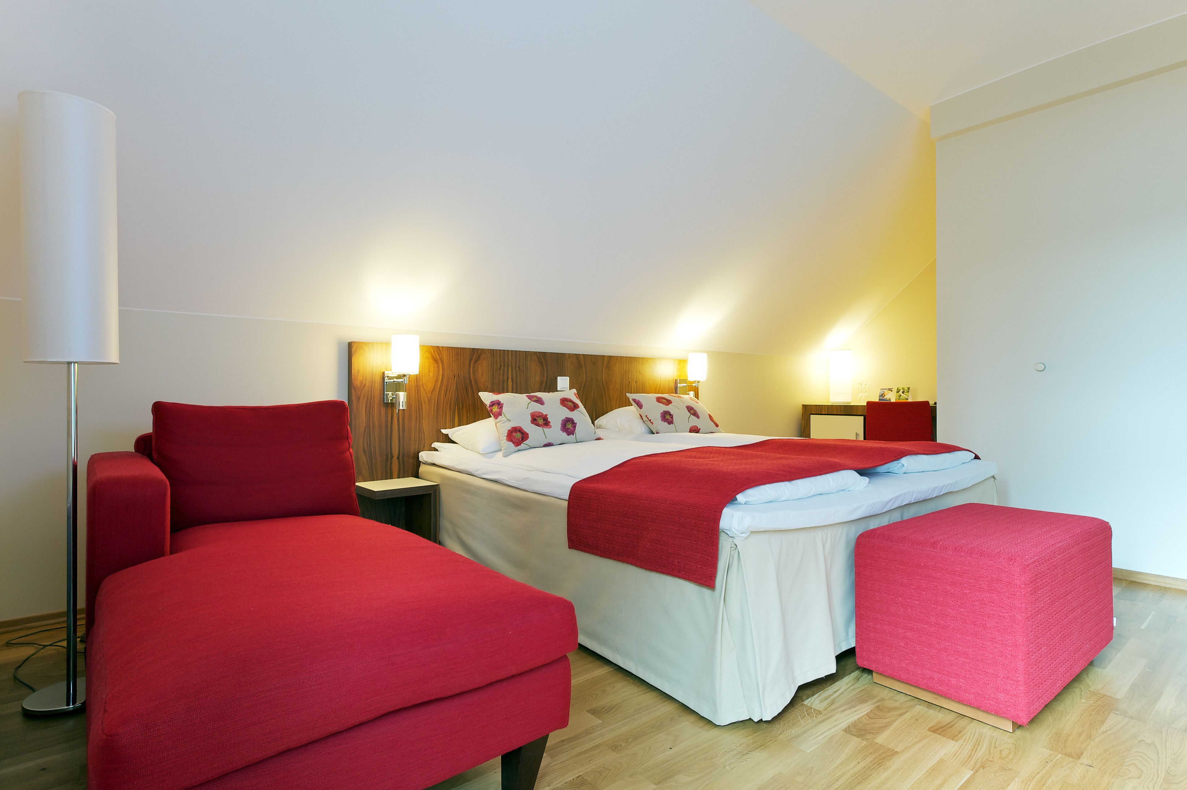 Una camera d'albergo con un letto, due divani rossi, un comodino e una lampada.