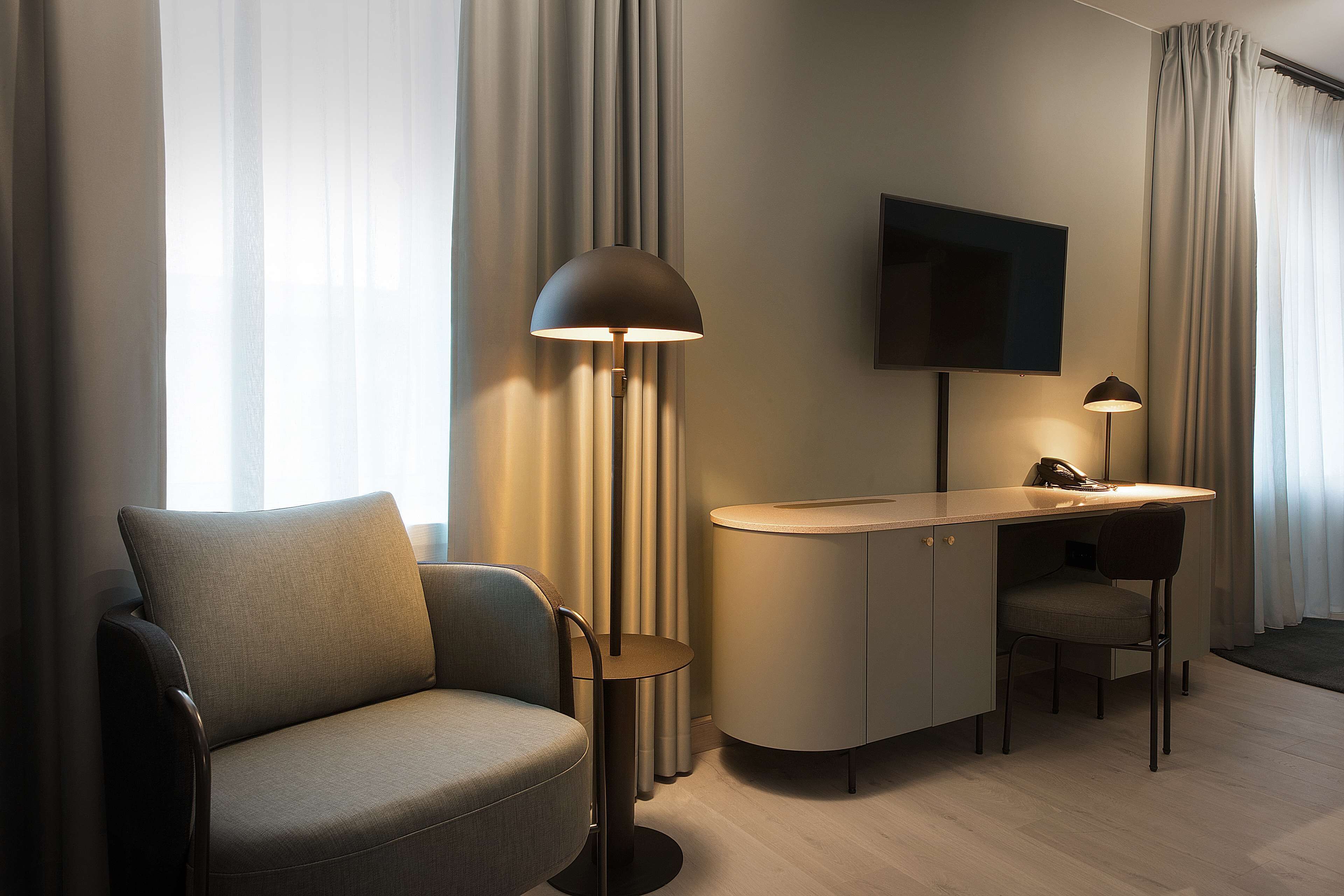 A modern hotel room with a sofa, a desk, a chair, a TV, and a floor lamp.