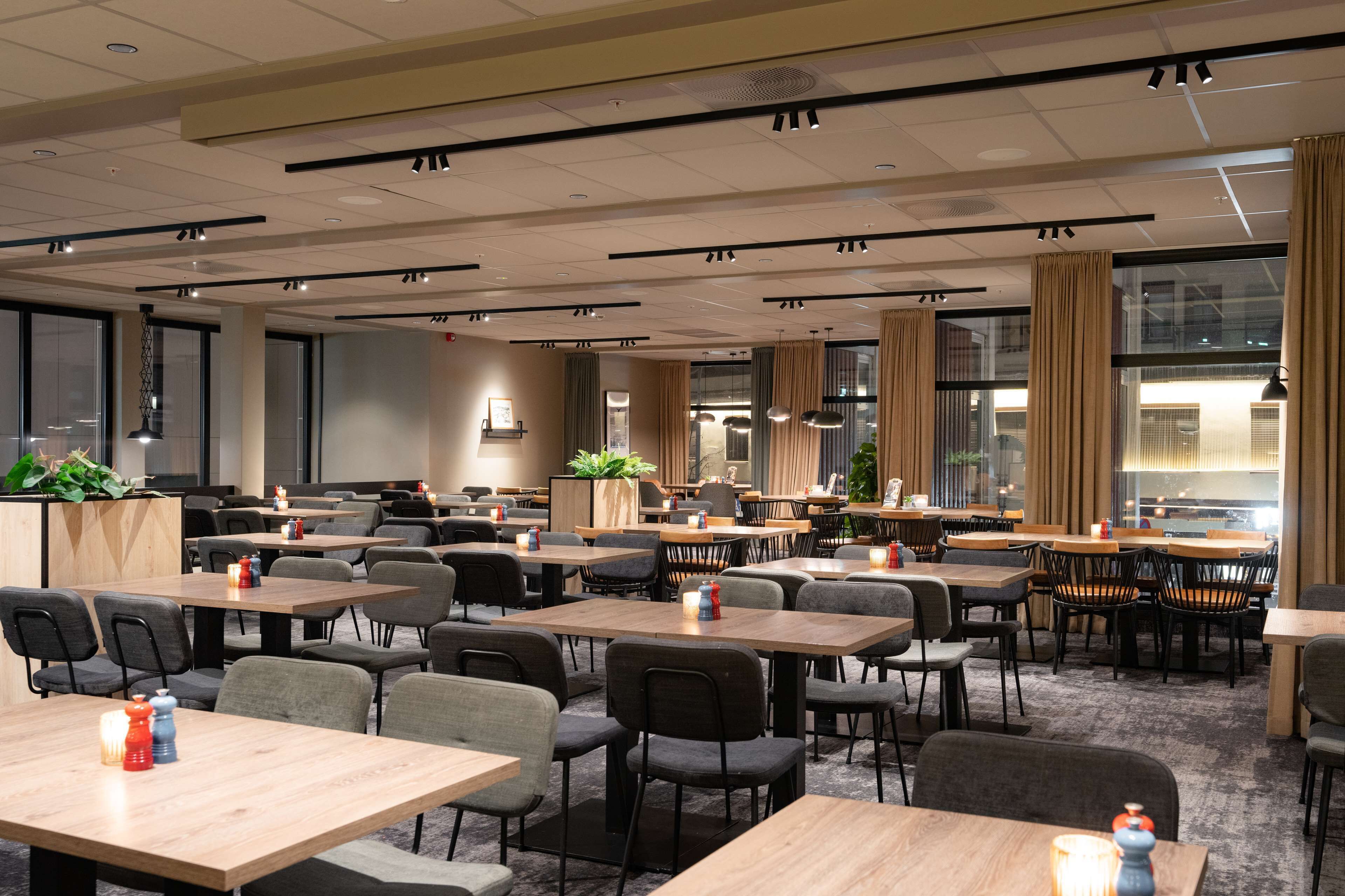 A hotel dining area with multiple tables and chairs, large windows with curtains, and ceiling lights.