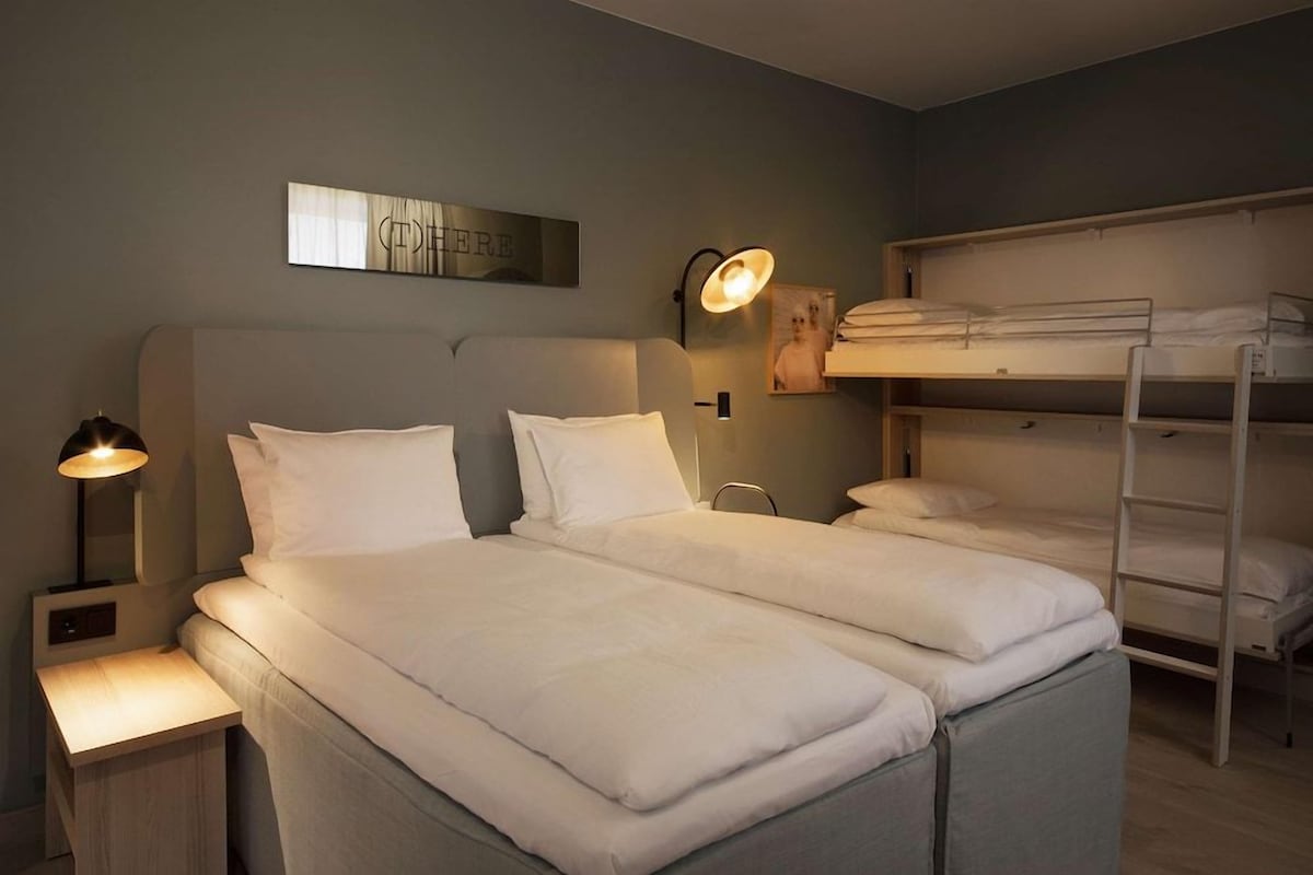 Un dormitorio con una cama, una mesita de noche, una lámpara de pared, una cama individual superpuesta y un cuadro en la pared.