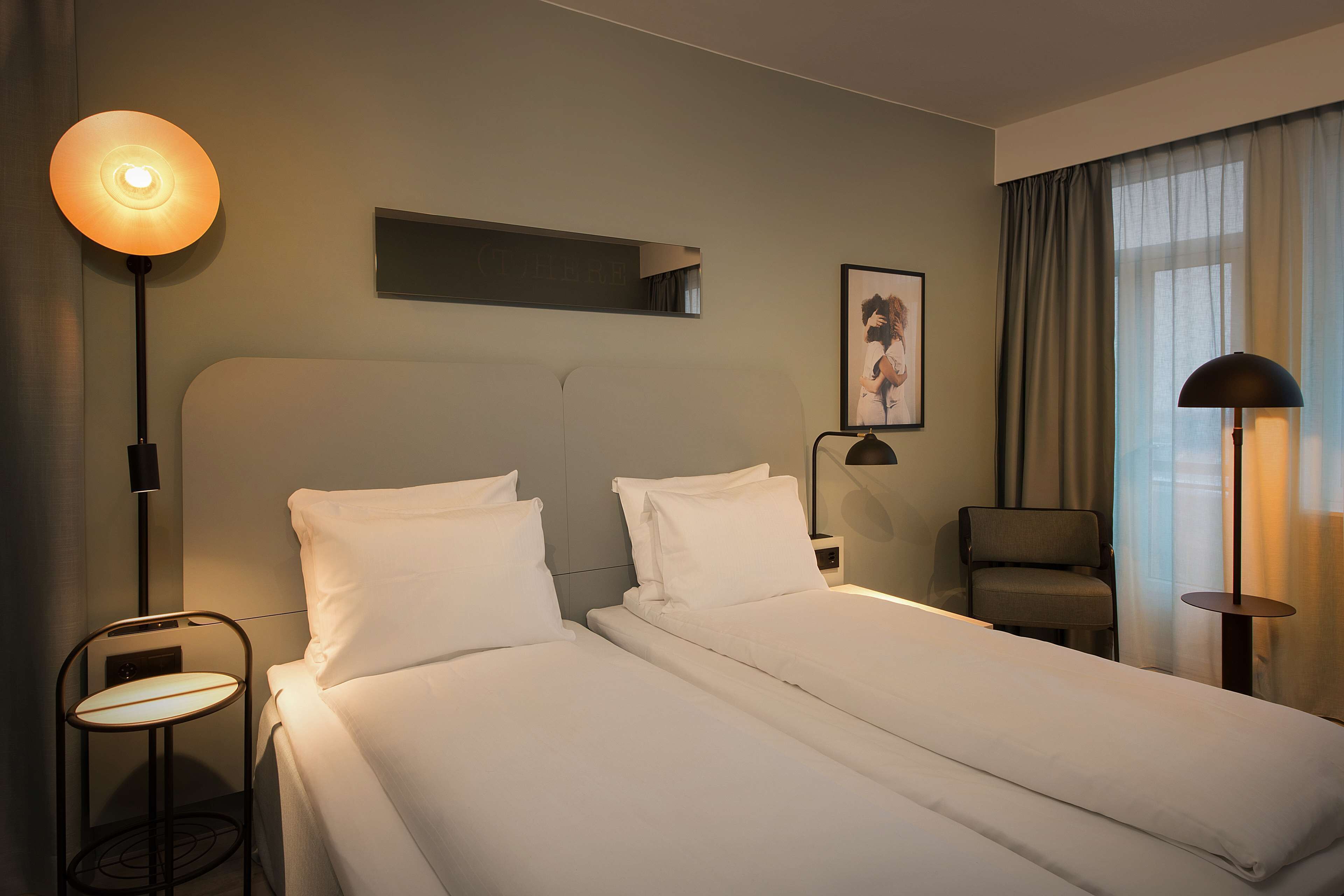 A hotel room with two single beds, a small table, a chair, a desk, and a large window with curtains.