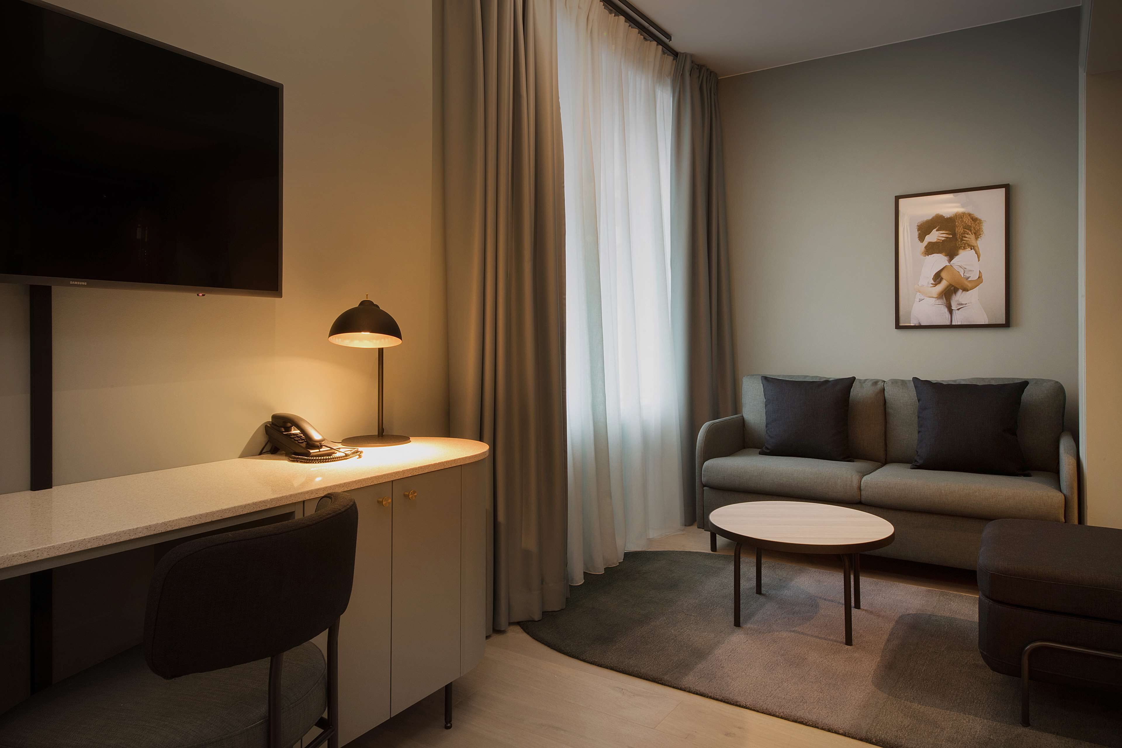 Habitación de hotel moderna con televisión de pantalla plana, escritorio con lámpara, sofá e imagen enmarcada en la pared.