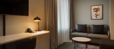Habitación de hotel moderna con televisión de pantalla plana, escritorio con lámpara, sofá e imagen enmarcada en la pared.