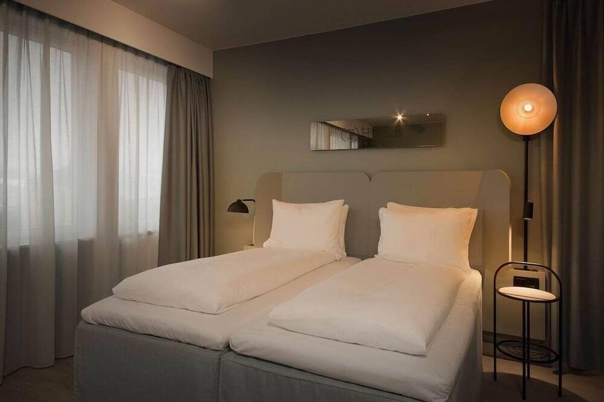 A double bed with white linens, a bedside table with a lamp, and a wall-mounted mirror.