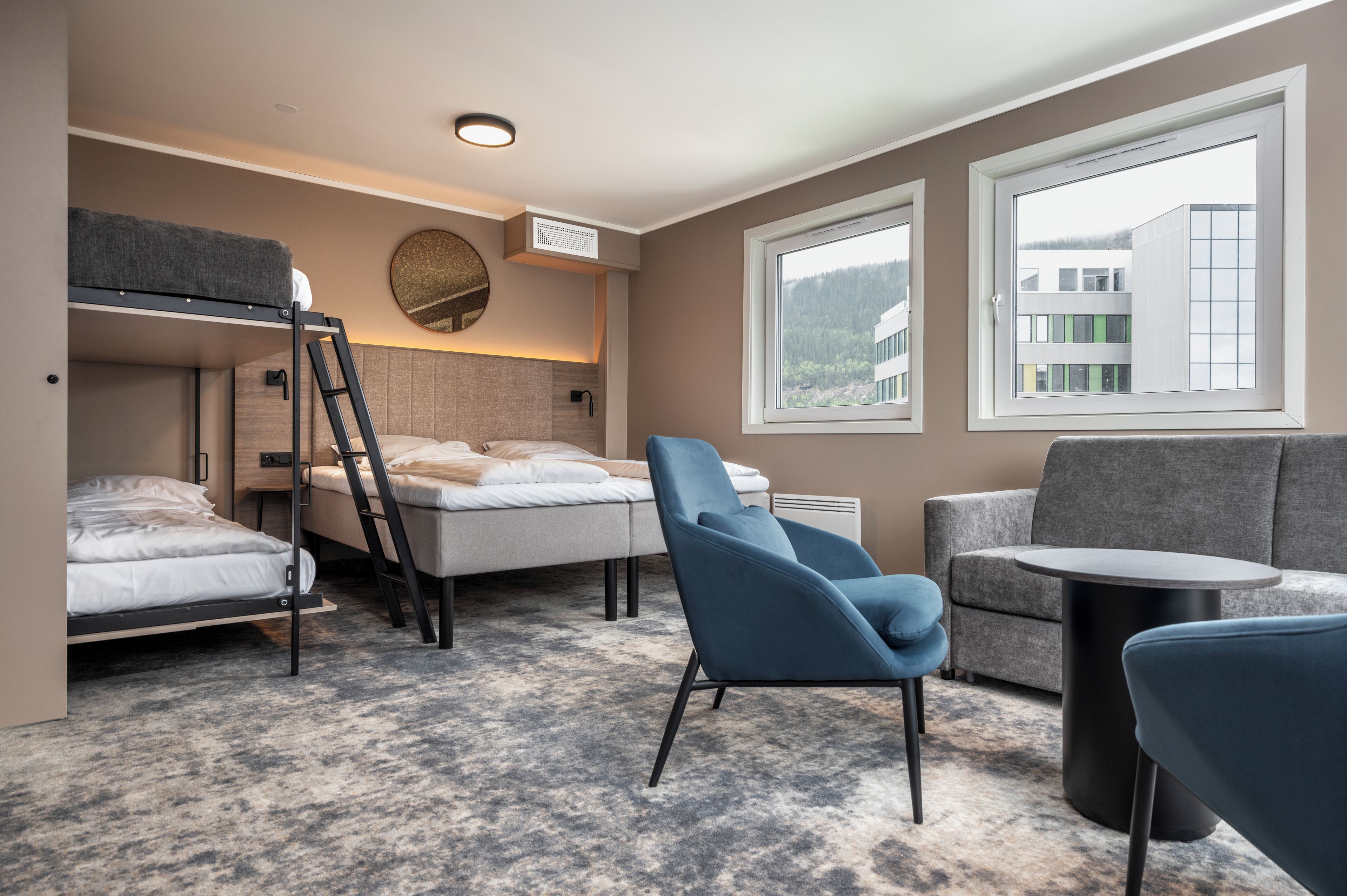 A compact hotel room with bunk beds, a desk, a chair, a round table, and large windows offering a view of buildings and trees.