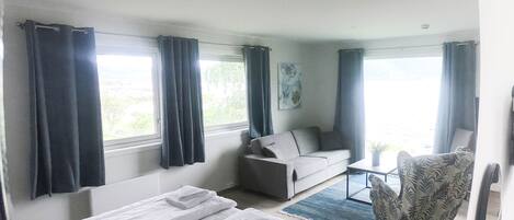 Quarto com cama, sofá, poltrona e uma mesa pequena. Janelas com cortinas.