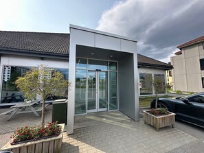 A modern building with a glass entrance, a paved courtyard, and a car parked nearby.